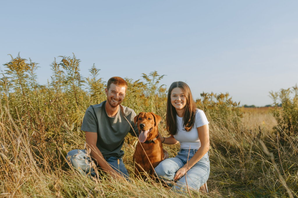 couple smiling at camera kneeled on ground posing with dog for engagement photos